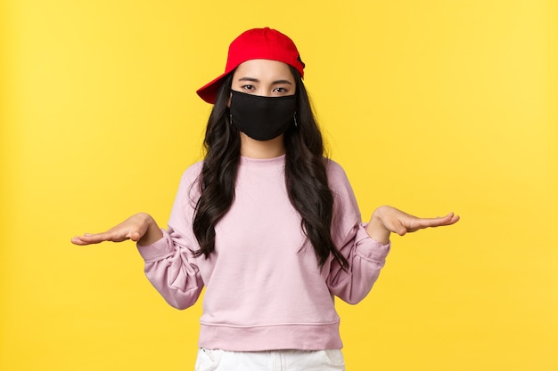 Covid-19, social-distancing lifestyle, prevent virus spread concept. unbothered young asian girl in face mask and red cap staying low, showing something little, yellow background.