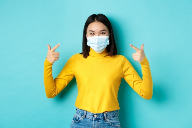 Covid-19, social distancing and pandemic concept. Young asian woman protect herself from coronavirus, pointing finger at her medical mask, standing over blue background