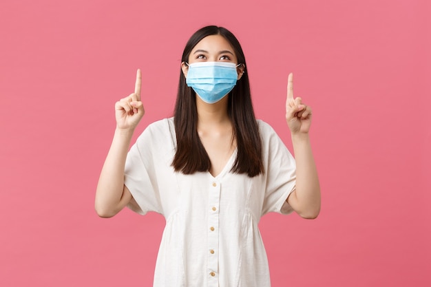 Covid-19, social distancing, virus and lifestyle concept. Delighted cute girl in medical mask and white dress, reading funny sign, pointing and looking up satisfied, smiling over pink background.