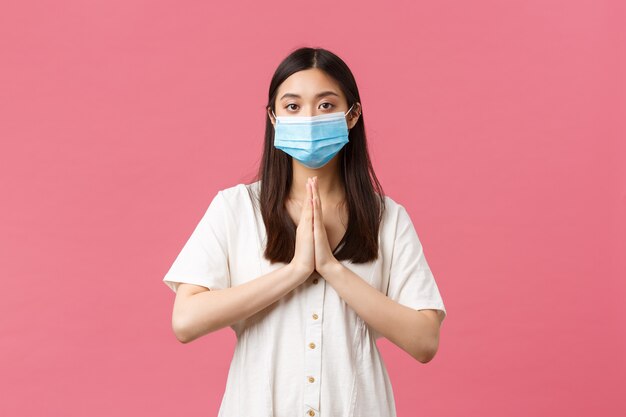 코비드-19, 사회적 거리, 바이러스 및 생활 방식 개념. 의료용 마스크를 쓴 희망찬 귀여운 아시아 소녀는 열정이 부족하고, 진지한 우울한 얼굴, 애원하는 분홍색 배경으로 도움을 요청하거나 호의를 구걸합니다.