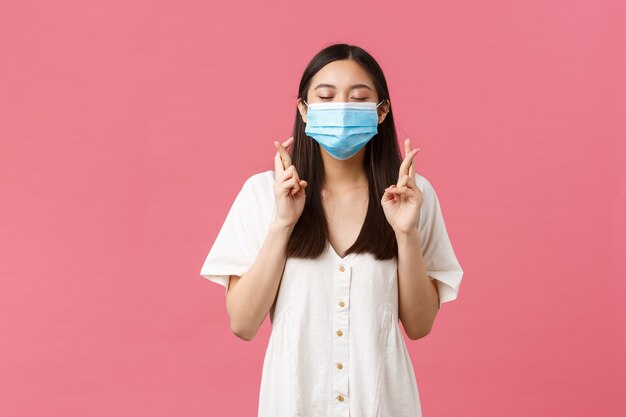 Covid-19, social distancing, virus and lifestyle concept. Hopeful dreamy asian girl making wish, close eyes and smiling in medical mask, cross fingers good luck, pleading over pink background.