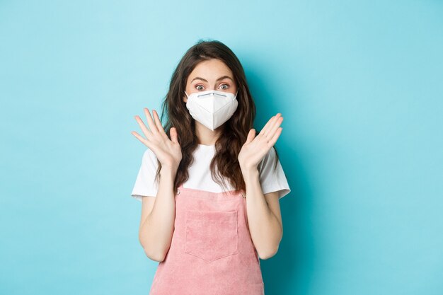 Covid-19, vaccination and quarantine concept. Excited and surprised young woman in medical respirator, face mask from coronavirus, clap hands and look amazed at camera, blue background