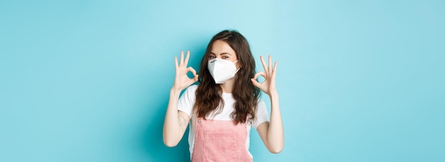 Бесплатное фото Концепция здоровья и пандемии covid очень хорошо молодая поддерживающая женщина, носящая медицинский респиратор и показывающая хорошие знаки одобрения, хвалит в масках для лица на общественном синем фоне