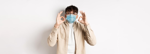 無料写真 covidの健康と実在の人々のコンセプトは、無菌のフェイスマスクを着て興奮し、感銘を受けた男で、okサインを示しています