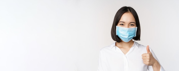 Free photo covid and healthcare concept portrait of asian woman wearing medical face mask and showing thumbs up standing over white background