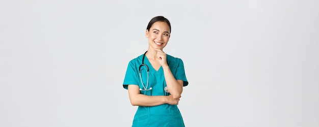 Free photo covid healthcare workers pandemic concept smiling pleased attractive asian female doctor in scrubs l...