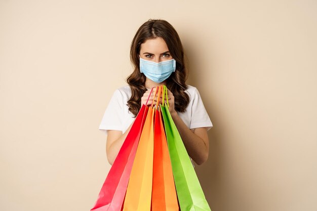 코비드 팬데믹과 라이프스타일 개념 젊은 여성이 의료용 얼굴 마스크를 쓰고 쇼핑백을 들고 포즈를 취하고 있습니다.