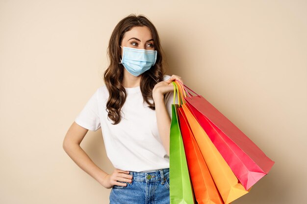 Covid pandemic and lifestyle concept young woman posing in medical face mask with shopping bags from...