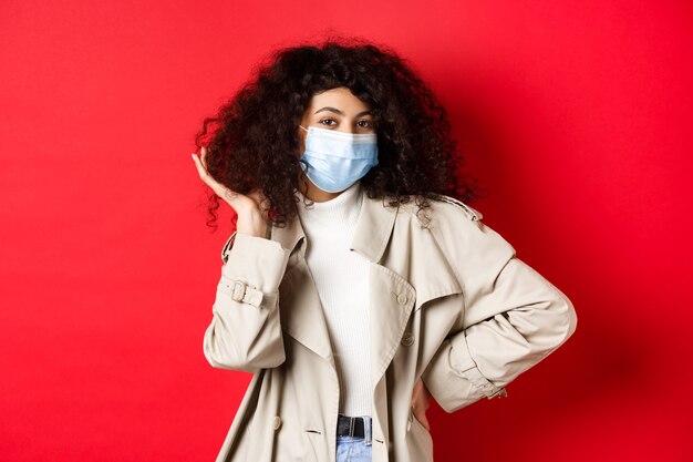 Covid pandemic and quarantine concept stylish coquettish woman in medical mask and trench coat fixin...