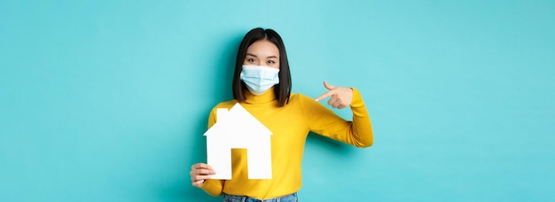 Free photo covid pandemic and real estate concept cheerful asian woman smiling in medical mask showing paper ho