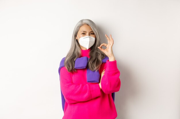 Covid, pandemic and social distancing concept. Cheerful and stylish asian senior woman wearing respirator from coronavirus, showing OK sign, standing over white background.