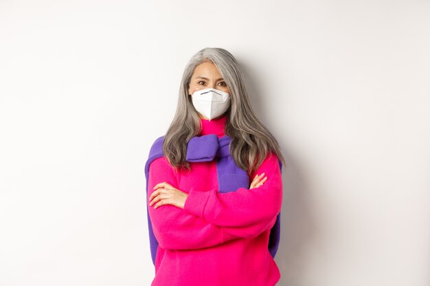 Covid, pandemic and social distancing concept. Determined and serious asian senior woman in face mask looking confident, cross arms on chest, standing over white background.