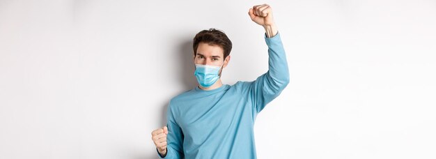 Foto gratuita pandemia di covid e concetto di allontanamento sociale ragazzo felice e fortunato in maschera medica che vince alzando han