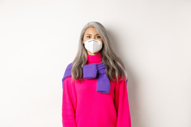 Covid, pandemic and social distancing concept. Stylish asian senior woman wearing respirator and looking at camera serious, standing over white background