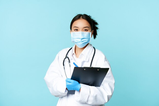 Covid and quarantine concept smiling asian female physician nurse in face mask holding clipboard shi...