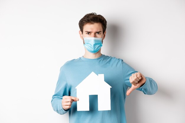 Covid and real estate concept. Disappointed young man in medical mask showing paper house cutout and thumb down, dislike broker agency, standing displeased over white background.