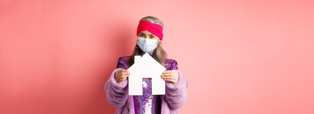 Free photo covid and real estate concept senior asian woman in stylish outfit and medical mask showing paper ho