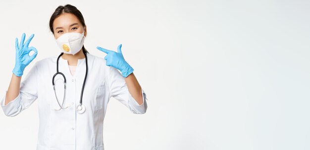Free photo covid and social distancing concept woman in medical respirator showing okay sign and pointing at he