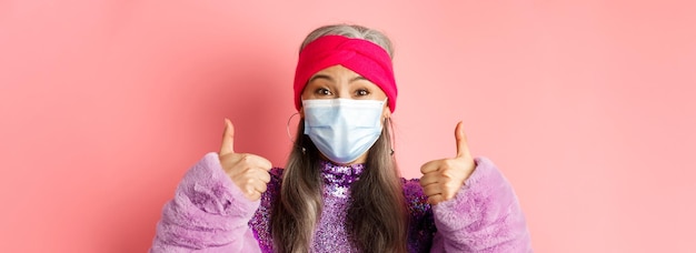 무료 사진 코비드 바이러스와 패션 컨셉은 엄지손가락을 보여주는 얼굴 마스크를 쓴 유행하는 아시아 노인 여성의 근접 촬영
