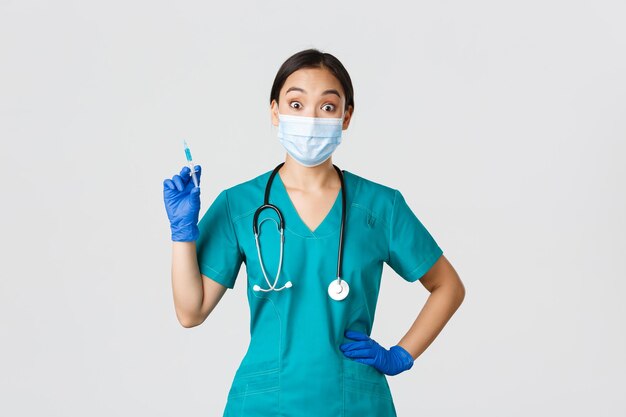 Covid19 coronavirus disease healthcare workers concept Amused smiling asian physician female doctor in medical mask and rubber gloves prepared syringe with vaccine for shot white background