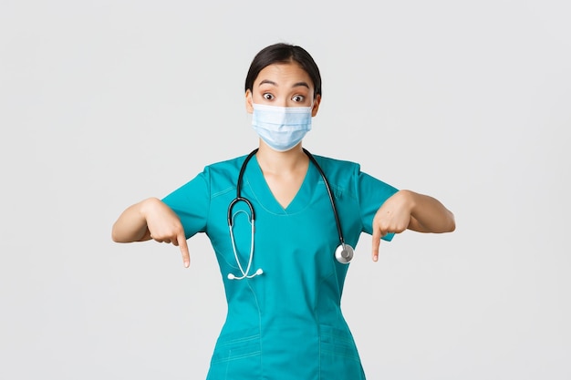 Covid19コロナウイルス病の医療従事者の概念スクラブと医療マスクで興奮して驚いたアジアの女性医師の看護師が広告を表示して指を下に向ける