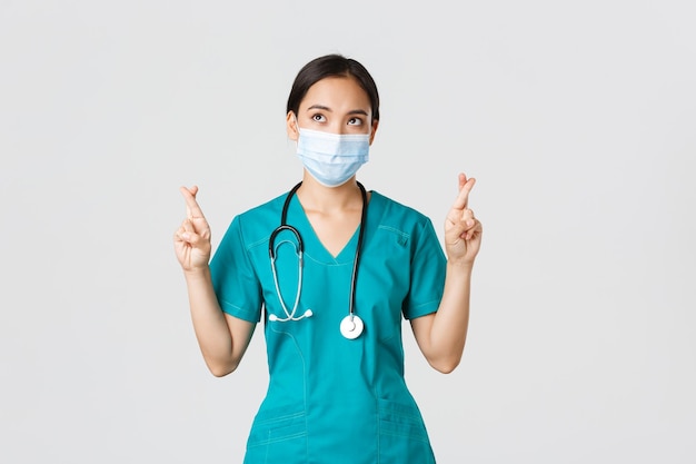 Covid19 coronavirus disease healthcare workers concept Hopeful seriouslooking asian doctor female physician in medical mask and scrubs cross fingers looking upper left corner