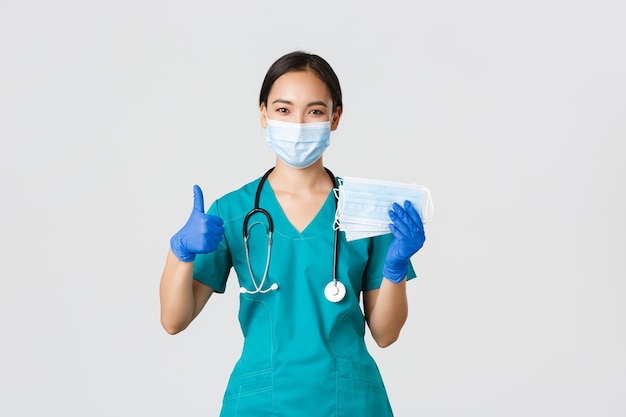 Концепция медицинских работников коронавирусной болезни Covid19 Улыбающаяся азиатская медсестра-врач в скрабах и резиновых перчатках, показывающая большой палец и медицинские маски на белом фоне