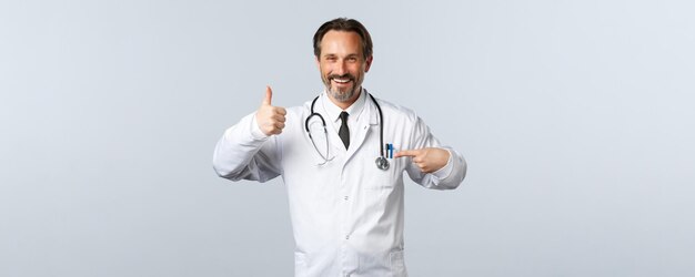 Covid19コロナウイルスの発生医療従事者とパンデミックの概念白衣を着た陽気な男性医師が製品を左に向け、親指を立てる承認は投薬または診療所を推奨します