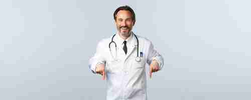 Free photo covid19 coronavirus outbreak healthcare workers and pandemic concept cheerful smiling male doctor in white coat inviting take test in clinic pointing fingers down advertising