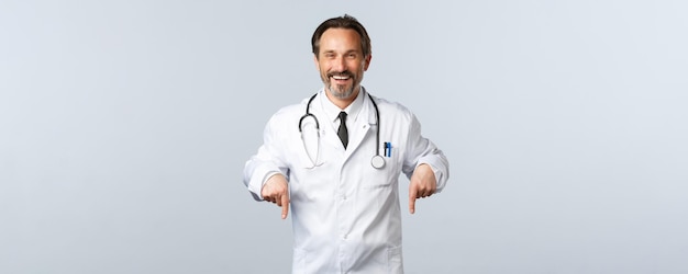 Covid19コロナウイルスの発生医療従事者とパンデミックの概念白衣を着た陽気な笑顔の男性医師がクリニックでテストを受け、指を下に向けて広告を出す