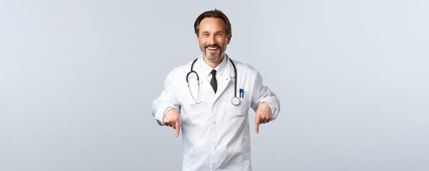 Covid19 코로나바이러스 발병 의료 종사자와 전염병 개념 흰색 코트를 입은 쾌활한 웃는 남성 의사가 광고를 가리키는 손가락을 가리키는 클리닉에서 시험을 봅니다.