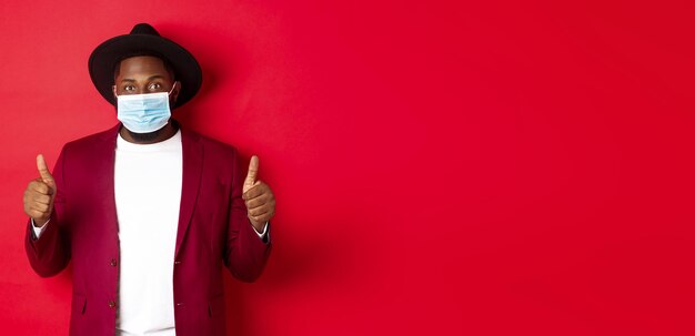 Free photo covid19 and fashion concept stylish african american man in hat and blazer wearing face mask and showing thumb up standing over red background