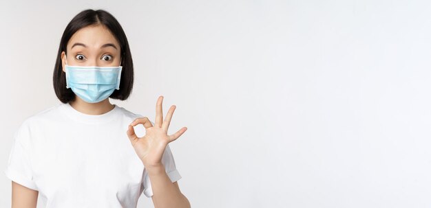 Covid19 healthcare and medical concept Impressed asian woman in medical mask looking amazed and showing ok okay sign standing over white background