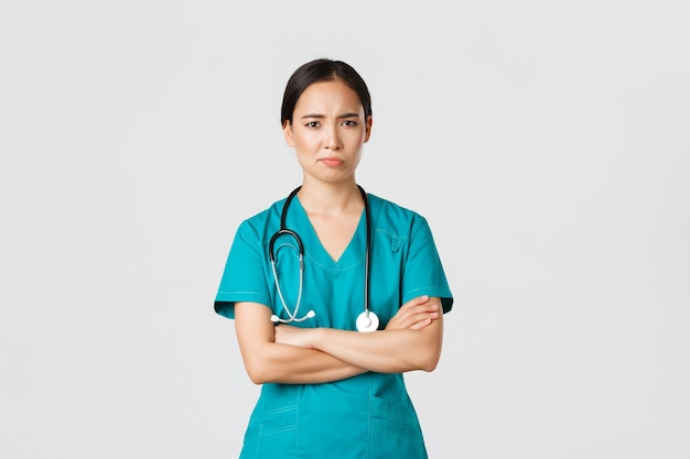 無料写真 covid19医療従事者のパンデミックの概念懐疑的で気が進まないアジアの女性医師は、スクラブクロスアームと眉をひそめている笑顔で疲れた看護師が立っている白い背景に不満を持っています