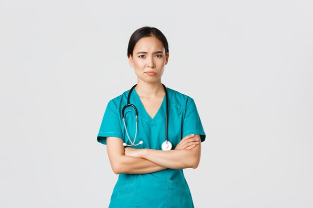 Covid19医療従事者のパンデミックの概念懐疑的で気が進まないアジアの女性医師は、スクラブクロスアームと眉をひそめている笑顔で疲れた看護師が立っている白い背景に不満を持っています