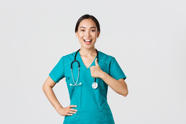 코비드19 의료 종사자의 대유행 개념 수술복을 입은 자신감 넘치는 미소 짓는 아시아 여성 간호사는 병원에서 서비스 품질을 보장하는 승인에 엄지손가락을 치켜들고 있습니다.