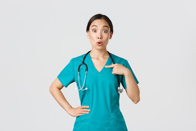 Covid19 healthcare workers pandemic concept Surprised and ambushed asian female physician nurse in scrubs pointing at herself beaing named or chosen standing white background