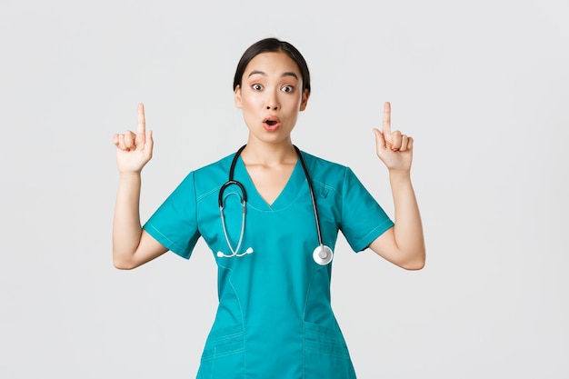 Covid19 концепция пандемии медицинских работников Удивленная и взволнованная азиатская женщина-медсестра женщина-врач в халате задает вопрос интересное промо, указывая пальцем вверх, показывая рекламу клиники