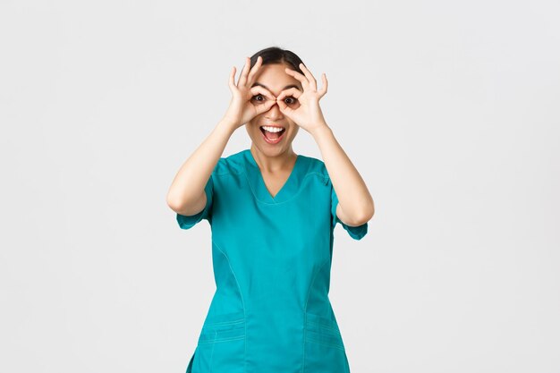 코비드19 의료 종사자와 바이러스 개념 예방 수술복을 입은 재미있고 귀여운 아시아 여성 간호사는 마치 쌍안경을 쳐다보는 것처럼 손가락으로 눈을 들여다보고 있습니다.