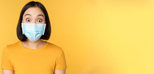 코비드19 및 의료 개념 얼굴 마스크를 쓴 아시아 여성의 초상화를 닫고 노란색 배경 위에 서 있는 뉴스에 놀라고 놀란 표정을 짓습니다