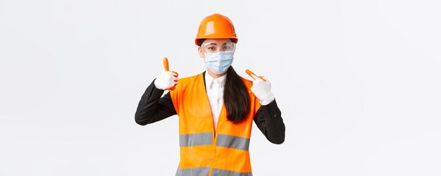 기업 건설 및 바이러스 개념 예방의 Covid19 안전 프로토콜 아시아 여성 수석 엔지니어는 직장에서 얼굴 마스크를 착용하고 그녀를 가리키고 승인에 엄지손가락을 보여줍니다