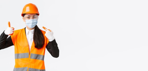 エンタープライズ建設とウイルスコンセプトの防止におけるCovid19安全プロトコルアジアの女性チーフエンジニアは、職場でフェイスマスクを着用して彼女を指さし、承認を得てサムズアップを示しています