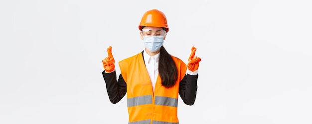 기업 건설 및 바이러스 개념 예방의 Covid19 안전 프로토콜 헬멧 안면 마스크를 쓴 희망적인 아시아 여성 엔지니어는 눈을 감고 손가락을 교차하여 행운을 빕니다.