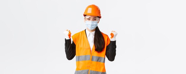 エンタープライズ建設でのCovid19安全プロトコルとウイルスの概念の防止フェイスマスクのヘルメットとメガネで成功した女性アジア人エンジニアが「はい」と言って拳ポンプで勝利