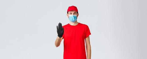 Covid19 selfquarantine online shopping concept Confident delivery man in red uniform gloves and face mask raise hand in stop sign prevent client step to storage without protective equipment