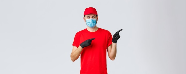 Covid19 selfquarantine online shopping and shipping concept Intrigued smiling delivery guy in red tshirt cap carrier uniform using medical mask and gloves to delivery to client point right