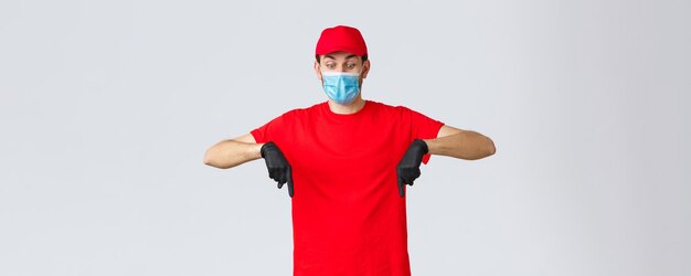 Covid19 концепция самокарантина онлайн-покупок и доставки Пораженный и шокированный курьер в униформе, красной кепке и футболке, в мерах безопасности, медицинской маске и перчатках, направленных вниз