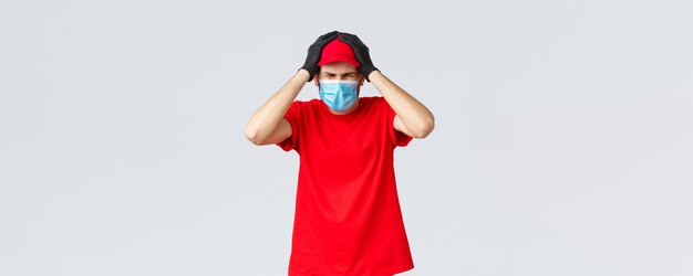 Covid19自己検疫オンラインショッピングと配送のコンセプト医療用マスクの手袋をはめた問題を抱えた恥ずかしい宅配便の男性が、小包の配達を間違えて顔をゆがめた頭に手をつないでいます