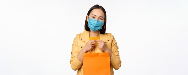 Covid19とショップのコンセプトパンデミックの白い背景の間にモールに行く買い物袋を保持している医療フェイスマスクを身に着けている若いアジアのスタイリッシュな女性