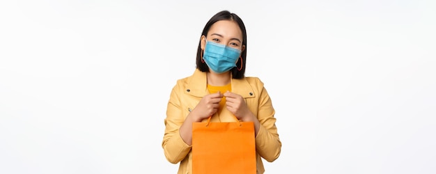 Covid19 and shop concept Young asian stylish woman wearing medical face mask holding shopping bag going in malls during pandemic white background